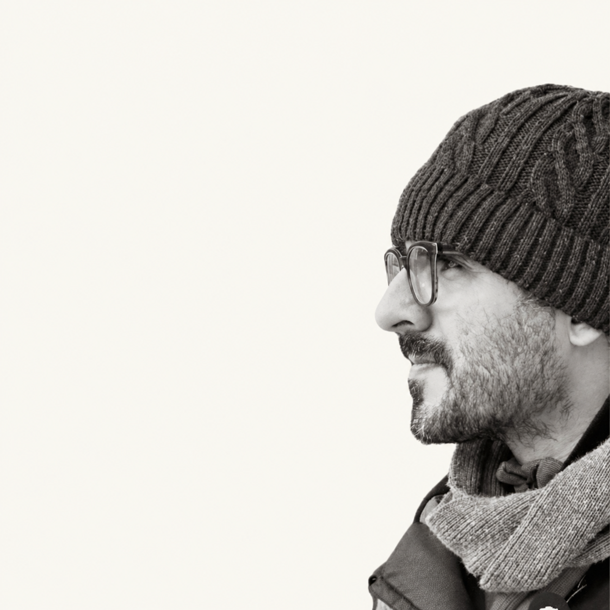 Ali Farjami in profile, wearing glasses and a knitted hat, against a warm ivory background